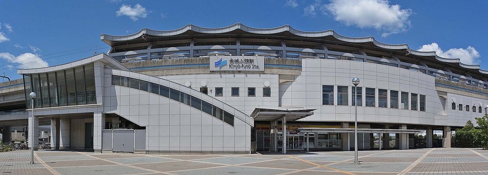 青波線金城ふ頭站的外觀寬幅照:白色高架站房頂上是波浪形的屋簷,正面掛著「金城ふ頭駅 Kinjō-futo Sta.」的站名招牌,左邊有一段斜向的玻璃樓梯通道,一樓有一間便利商店,站前是鋪著彩色地磚的空曠廣場