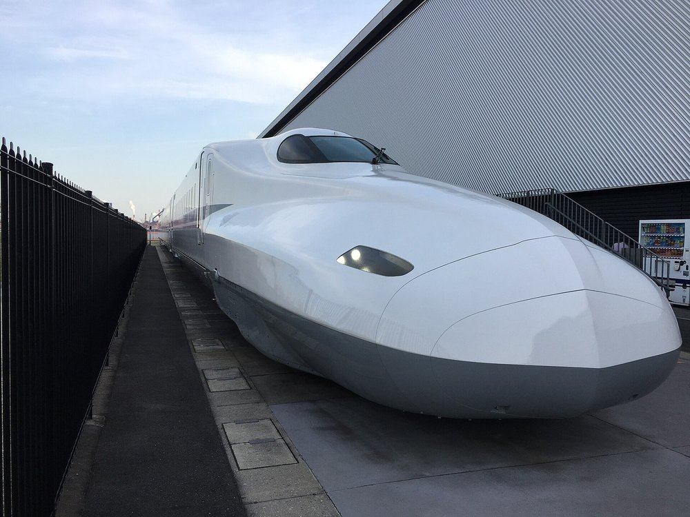 停在館外的一節白色 N700 系新幹線車頭,流線形的長鼻子朝向鏡頭,車身下半是灰色,左邊是黑色鐵柵欄和一條步道,右邊是館的灰色波浪板外牆、一道金屬樓梯和一台飲料自動販賣機