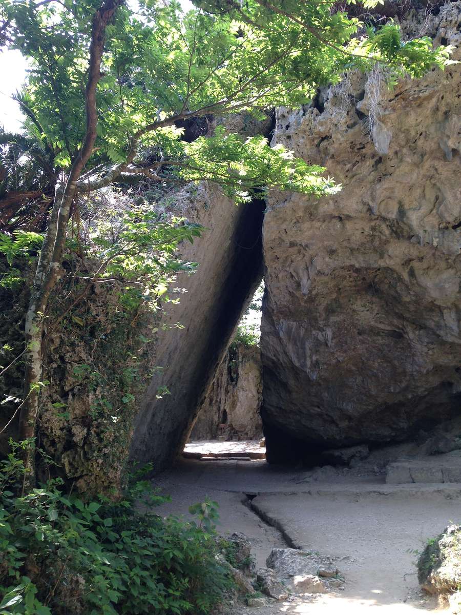 斎場御嶽的三庫理：兩塊巨大的石灰岩斜靠在一起，中間形成一道三角形的通道，地面是碎石步道，左側有樹枝與蘇鐵葉垂下
