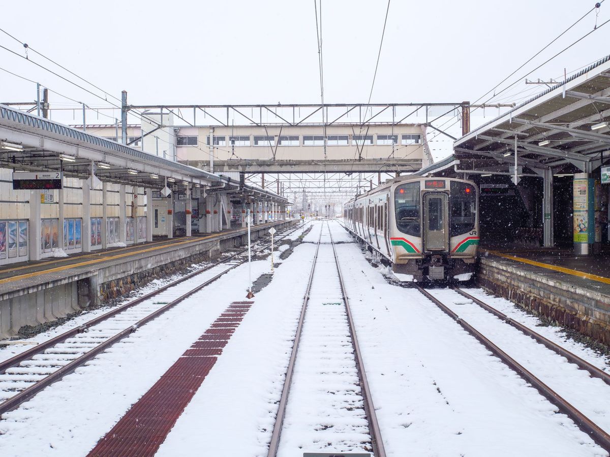 下雪中的會津若松站：月台之間的軌道積滿了雪，右側月台邊停著一列紅綠色塗裝、車頭有 JR 標誌的 E721 系電車，月台上沒有人