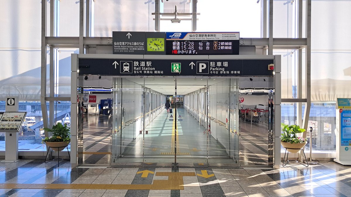 航廈 2 樓往車站的入口：一道玻璃門通進一條兩側全是玻璃的長廊，門上方黑底看板寫著「鉄道駅 Rail Station」和「駐車場 Parking」，最上面掛著仙台空港アクセス線のりば的指標、綠色時鐘與發車時刻板，地上有黃色導盲磚與箭頭，門兩側各擺一盆綠色植物