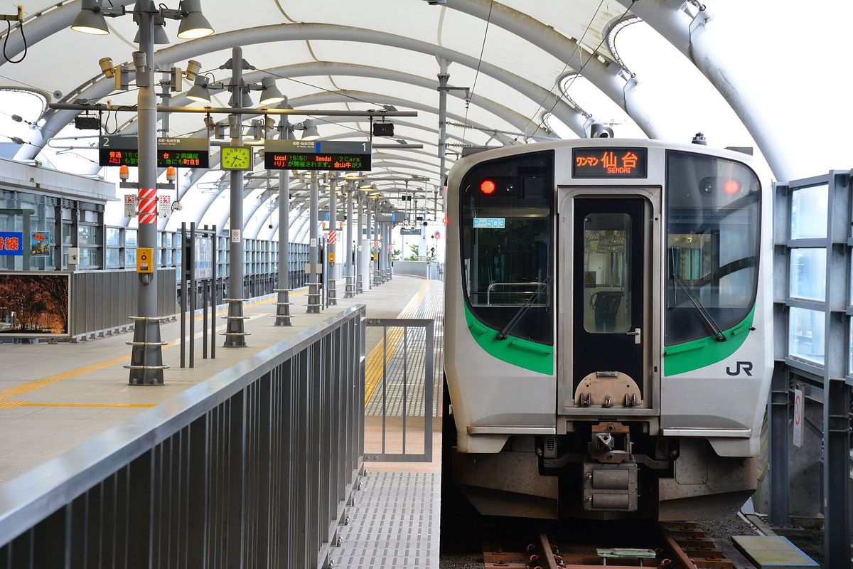 仙台空港站月台上停著一列 JR 東日本 E721 系列車，白色車頭帶綠色腰帶、右下角印著 JR 標誌，車頭上方的橘色字幕寫著「ワンマン 仙台 SENDAI」，左邊月台頂棚下掛著發車資訊板與綠色時鐘