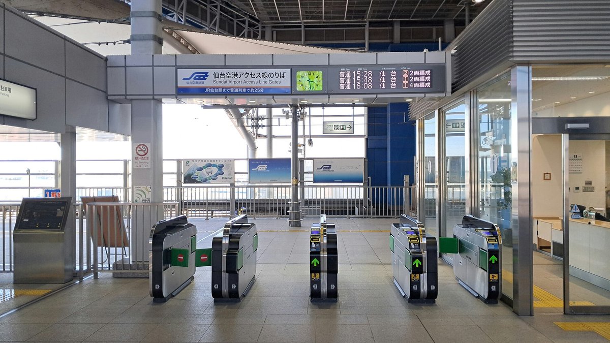 仙台空港站驗票口：一排五台黑色自動驗票機，幾台亮著綠色箭頭，上方看板寫著「仙台空港アクセス線のりば Sendai Airport Access Line Gates」與「JR仙台駅まで普通列車で25分」，旁邊是綠色時鐘和顯示 15:28、15:48、16:08 三班往仙台的發車板，右側是站務室窗口