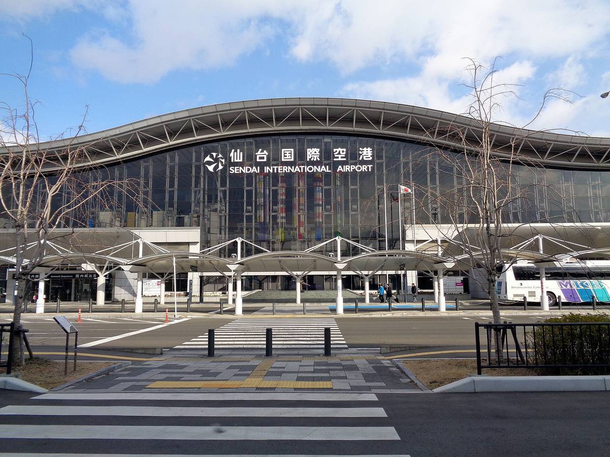從停車場那一側正面看仙台機場航廈：波浪形的銀色屋頂下是整面玻璃帷幕，上面掛著白字「仙台国際空港 SENDAI INTERNATIONAL AIRPORT」與機場標誌，一樓前方是一排白色傘狀遮雨棚，右邊停著一輛遊覽車，前景是斑馬線與兩棵沒有葉子的樹