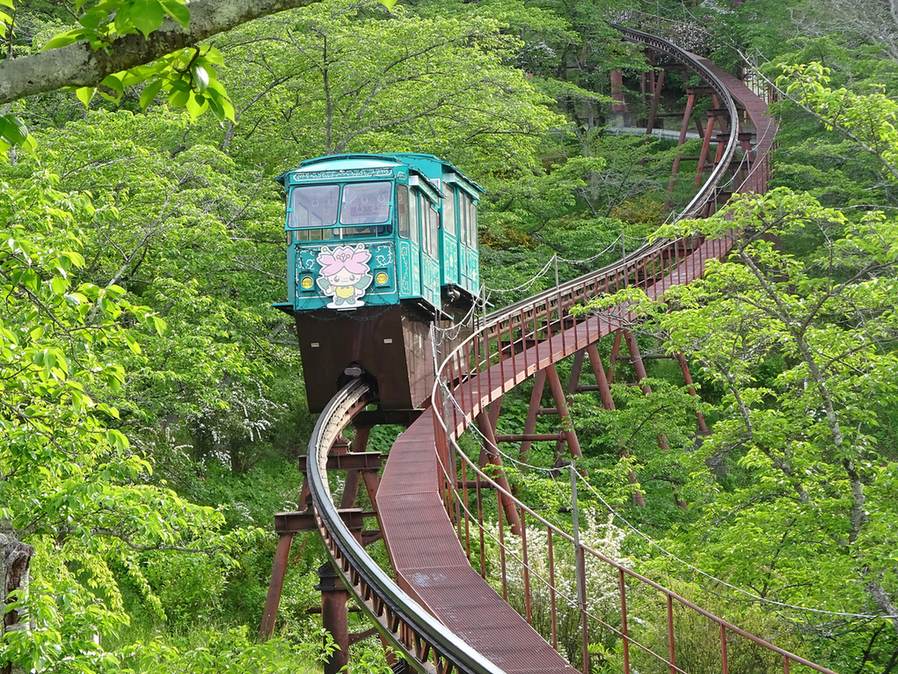 船岡城址公園的斜行電梯:一台綠色單軌小車停在架高的彎曲軌道上,車頭貼著櫻花造型的吉祥物,軌道旁邊有一條紅褐色的金屬步道,四周的櫻樹全是綠葉