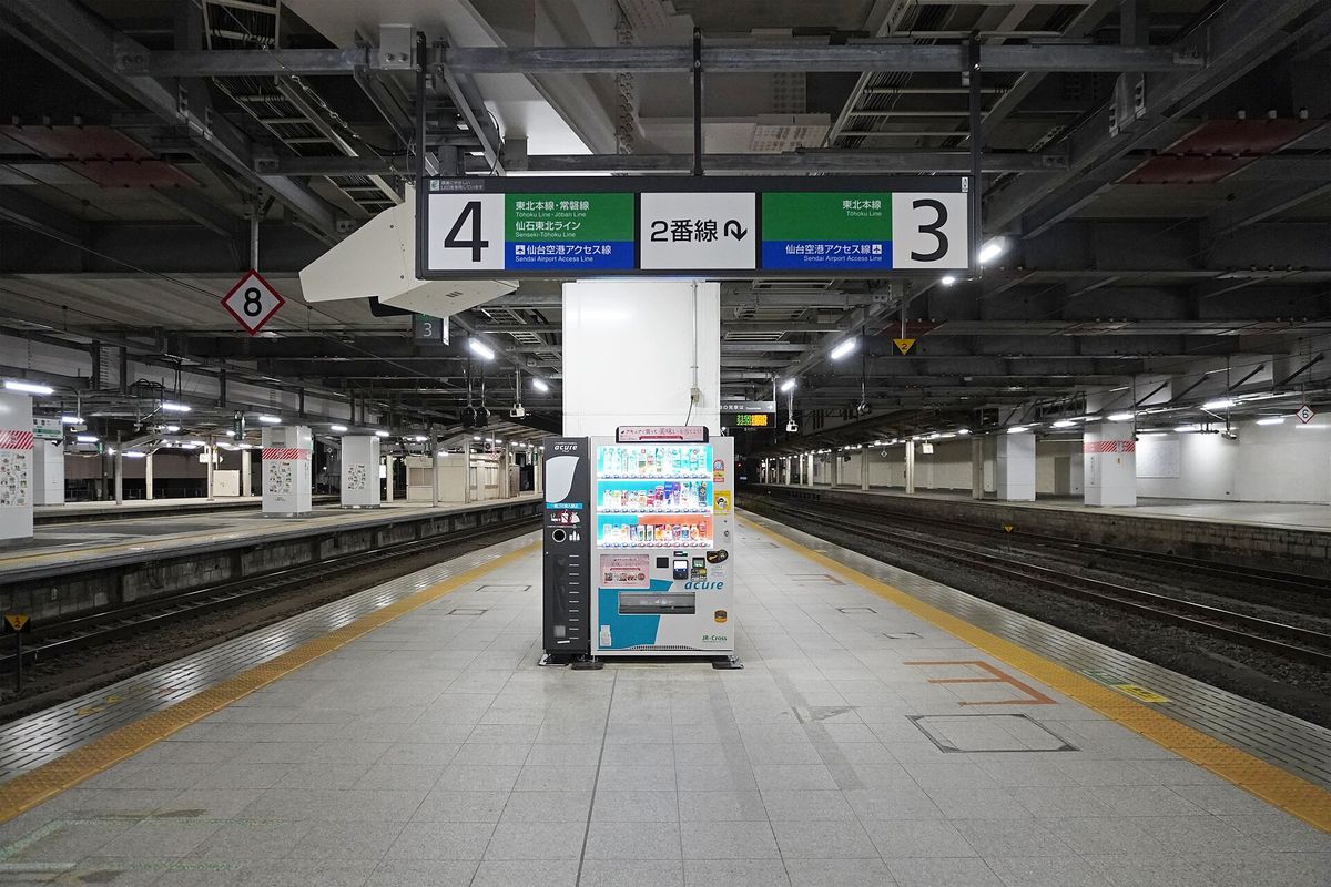JR 仙台站地面層的月台，中間柱子上方掛著站名板：左邊「4」番線寫著東北本線・常磐線、仙石東北ライン、仙台空港アクセス線，右邊「3」番線寫著東北本線、仙台空港アクセス線，中間是「2番線」的方向指示，柱子下方是一台自動販賣機，兩側軌道上沒有列車