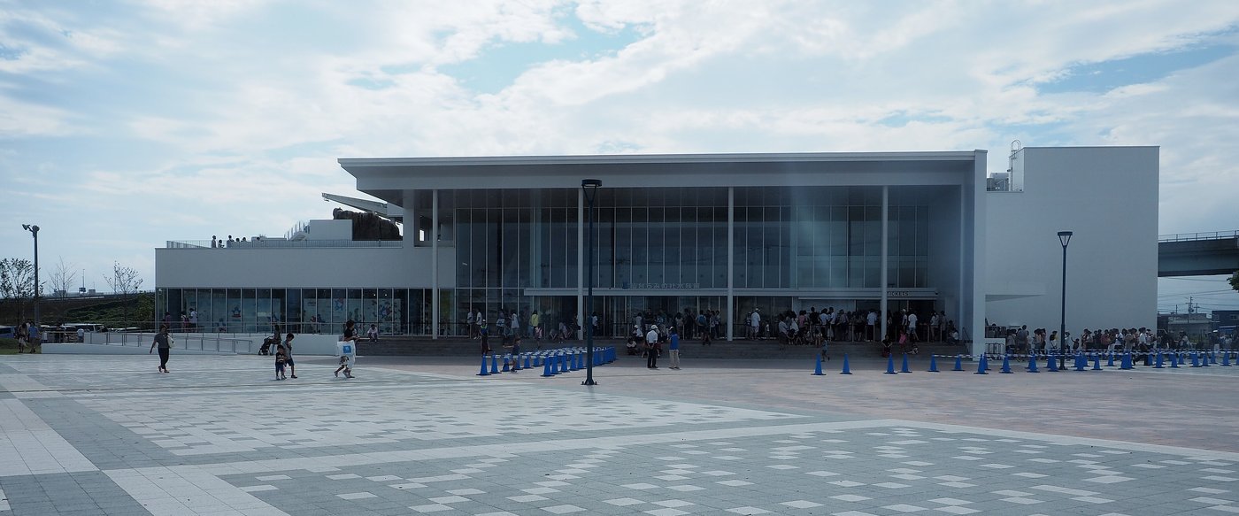 仙台海洋森林水族館外觀:白色方盒狀建築,正面是兩層樓高的整面玻璃帷幕,前方是大片灰白格紋的石板廣場,入口前有藍色交通錐圍出的排隊動線與許多遊客