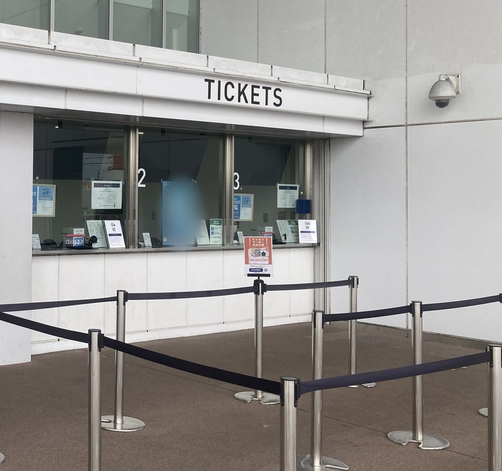水族館外牆上的售票窗口,白色雨庇上方寫黑字「TICKETS」,下方是編號 2、3 的玻璃窗口,窗內貼著幾張告示,窗口前用不鏽鋼立柱和黑色拉帶圍出排隊動線,沒有人在排