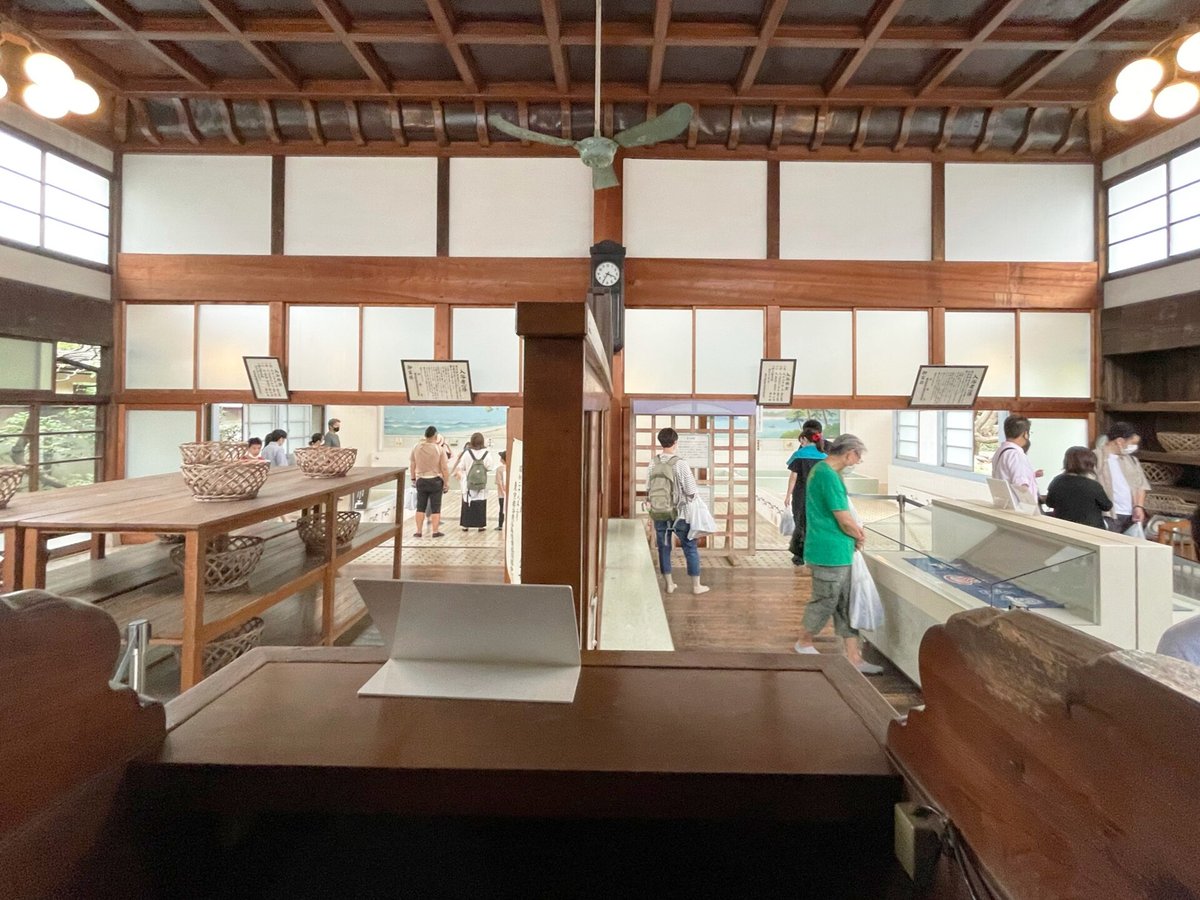 The view out from the attendant's seat at a bathhouse bandai: the wooden counter in the foreground, the men's and women's changing rooms opening to either side of a central pillar, wicker undressing baskets on shelves, and framed bather notices and a clock on the walls