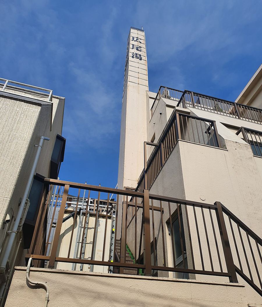 A tall square concrete bathhouse chimney in a back street in Hiroo, Shibuya, Tokyo, with the name Hiroo-yu written vertically down it against a blue sky, flanked by apartment blocks