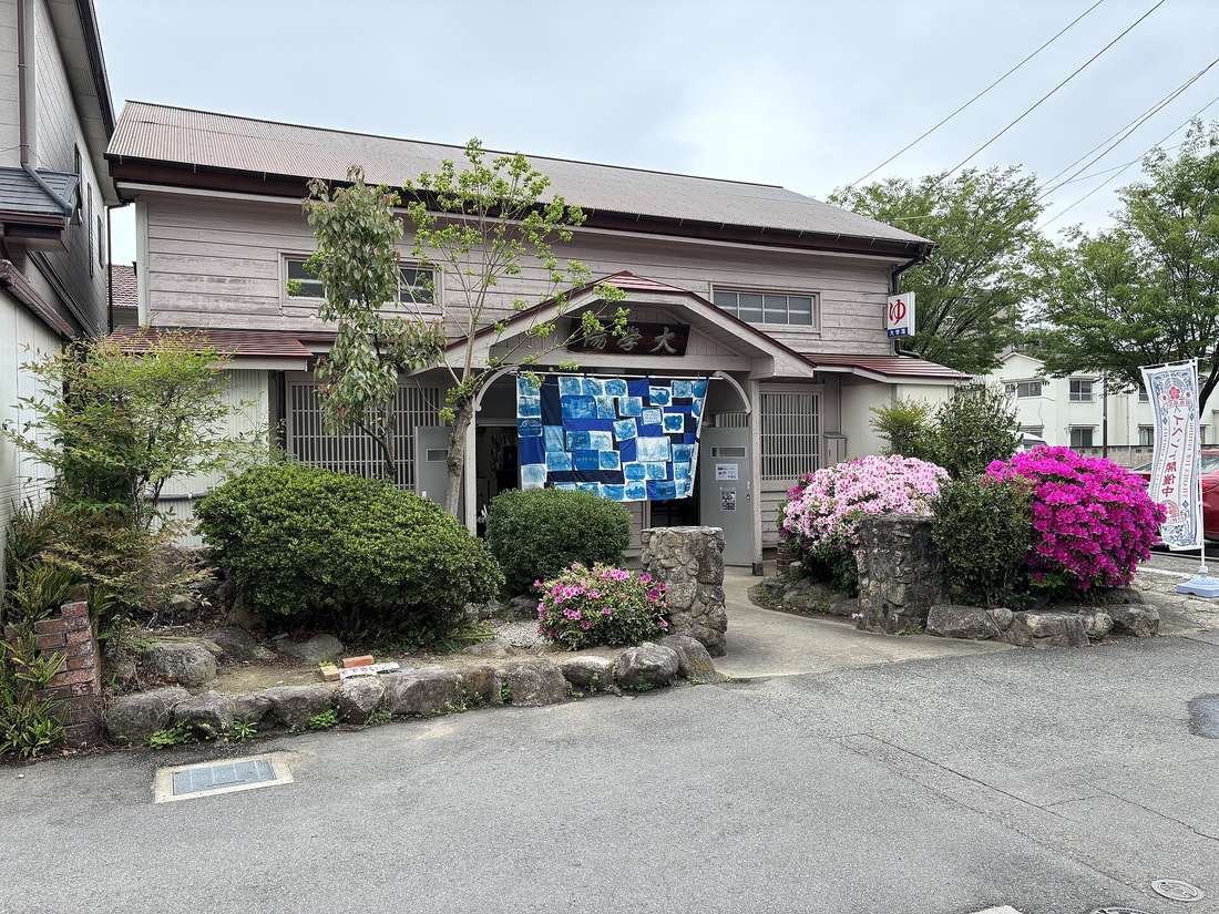 The wooden exterior of a Japanese public bathhouse, with an indigo noren curtain over the entrance and a yu character sign on the side wall