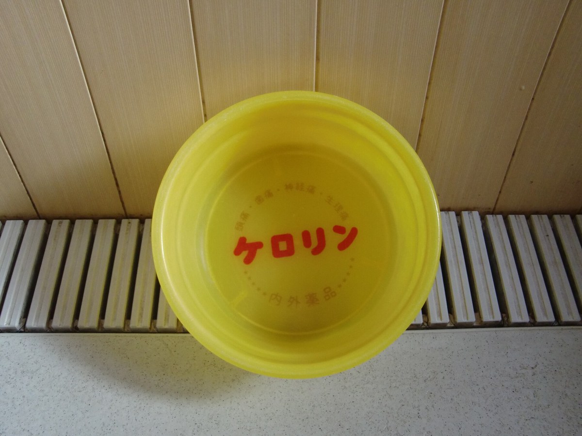 Overhead close-up of a single yellow Kerorin plastic wash basin, the brand name printed in large red characters in the base and the maker Naigai Yakuhin in smaller orange type around it