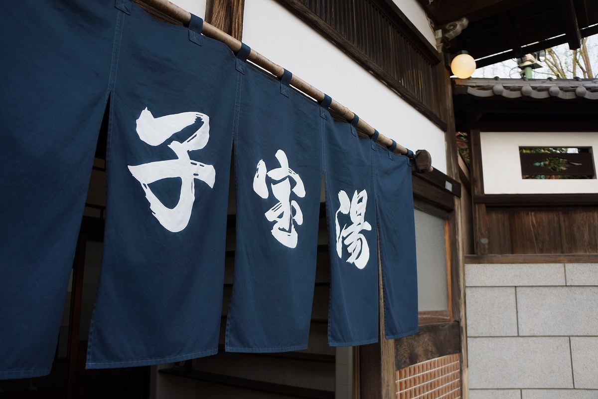 Close view of the indigo noren curtain at the entrance of the bathhouse Kodakara-yu, three white characters dyed on deep blue cloth, hung from a bamboo pole under old timber eaves