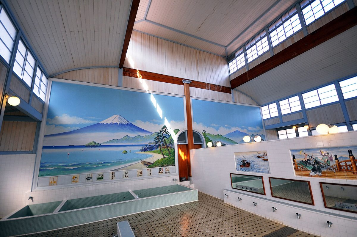 The full bath hall of a Japanese bathhouse: a painted mural of Mount Fuji and a coastline across the far wall, tiled rectangular soaking tubs to the left, and a row of taps and mirrors set low along the right-hand wall above octagonal floor tiles