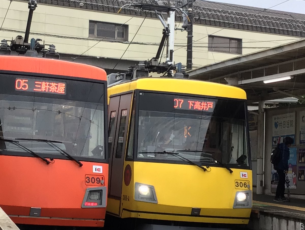 上町站並排停著兩輛東急世田谷線電車的車頭:左邊橘色車的顯示器寫「三軒茶屋」,右邊黃色車寫「下高井戸」,兩輛車頭都有 IC 標誌,右側月台上一位背對鏡頭的乘客