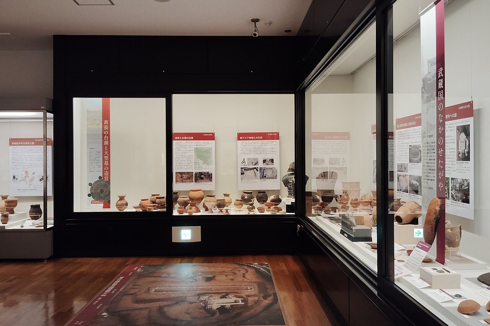 世田谷區立鄉土資料館的展示室:深色牆面嵌著玻璃展示櫃,櫃內排著成列的陶器與紅色標題的解說板,右側直幅標題寫著「武蔵国のなかのせたがや」,木地板上有一幅遺跡的空拍照