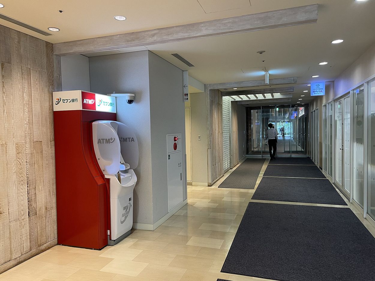 A red Seven Bank ATM standing in a Japanese building passageway, with Seven Bank and ATM signage above the machine