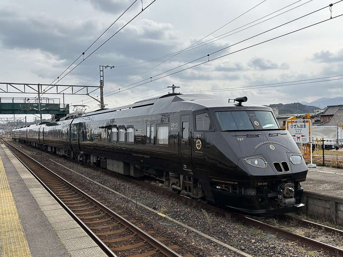 停在肥前浜站月台的 36ぷらす3:整列漆成黑色、車頭圓弧的電車,車身印著金色的 36+3 標誌,右邊月台上立著寫著ひぜんはま的站名牌,遠處是低矮的山