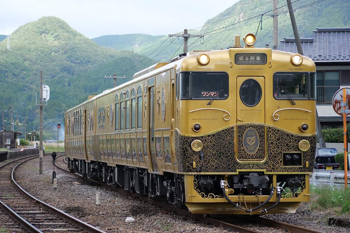 或る列車行駛在山間的單線鐵道上:金色車身的兩節柴油車,車頭下半部是黑底金色的唐草花紋,正中央一枚心形徽章,車頭上方的牌子寫著「或る列車 JRKYUSHU SWEET TRAIN」,背景是長滿樹的綠色山坡