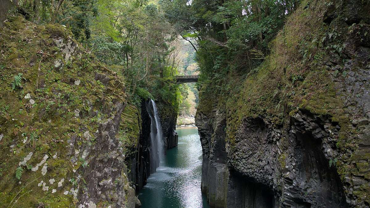 高千穗峽:兩側長滿青苔的柱狀岩壁夾著一條碧綠色的河,左側岩壁上一道細長的瀑布落進河裡,遠方有一座石造拱橋跨過峽谷