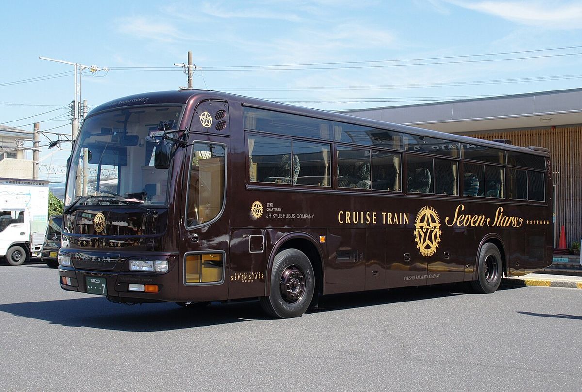 停在車站前廣場的深褐色大型遊覽巴士,車身側面用金色字寫著 CRUISE TRAIN 和 Seven Stars,中間是金色星形徽章,車門旁印著 CHARTERED JR KYUSHUBUS COMPANY