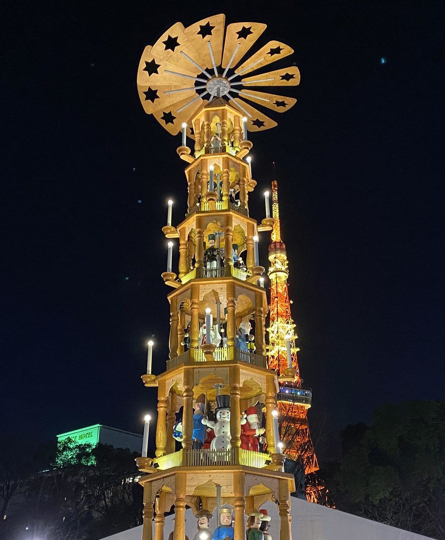 2019 年 12 月芝公園會場的聖誕金字塔直幅夜景:塔頂是鏤空星形的木製扇葉,往下五層各站著雪人、聖誕老人、士兵等木偶,每層外圈有燭形燈;右後方是點著橘色燈的東京鐵塔,左下角有一棟打綠光的飯店