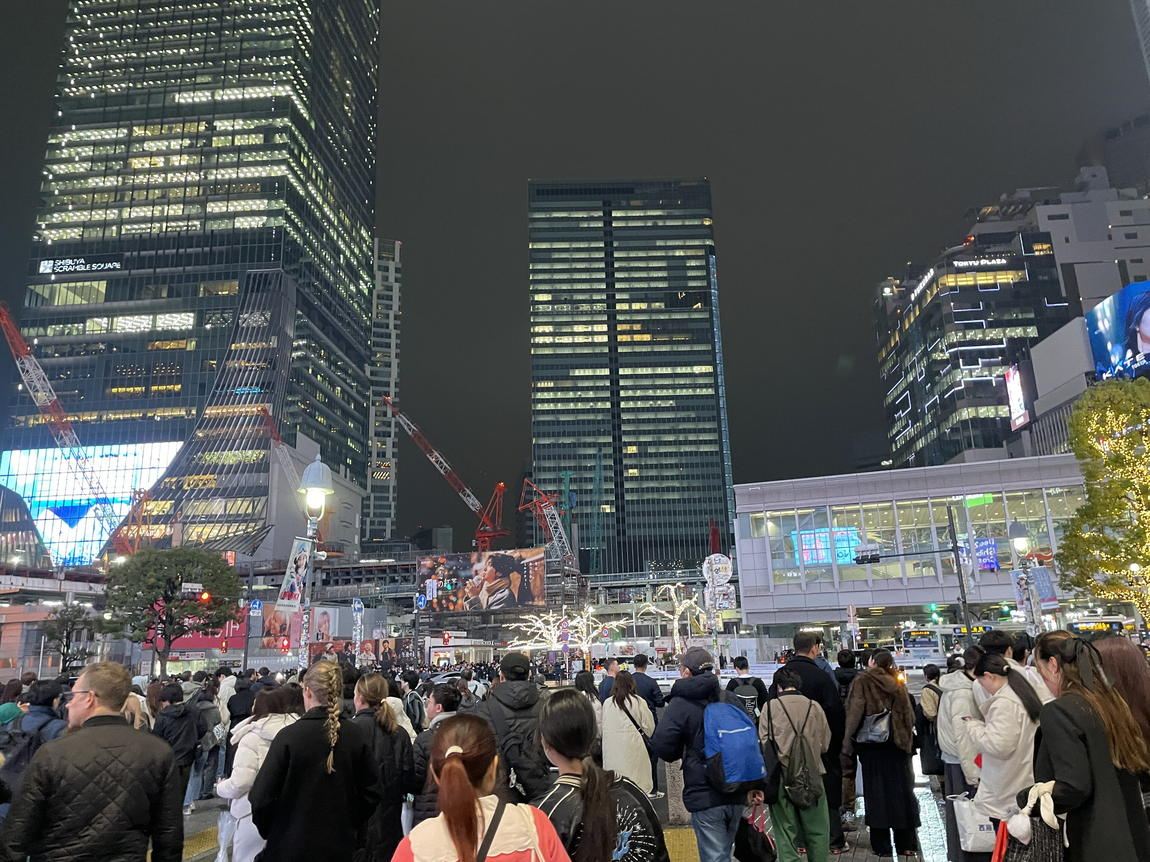 A dense crowd in dark winter coats and puffer jackets waiting to cross at Shibuya Crossing on Christmas Eve night, with Shibuya Scramble Square and Shibuya Station lit behind them