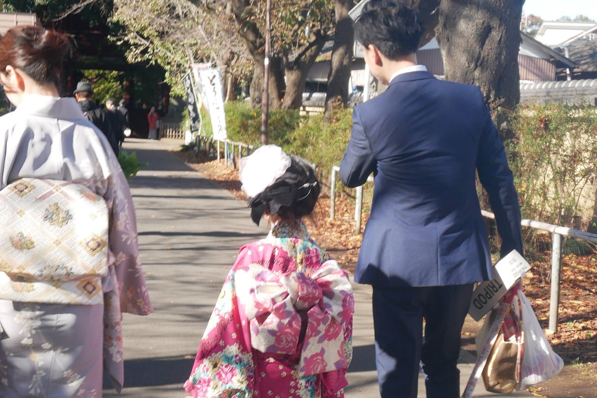 A girl in formal Shichi-Go-San kimono walking a shrine approach with both parents, all three seen from behind