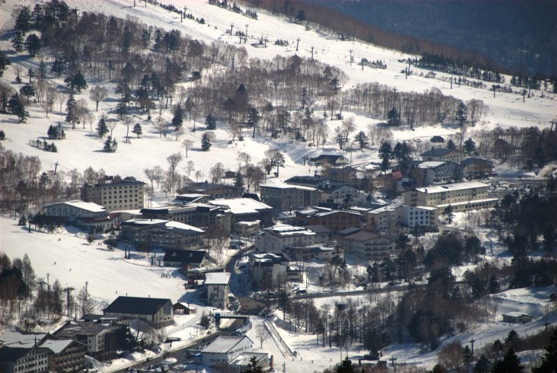 雪地裡密集的一片旅館與山莊建築，四周是落葉樹，後方的雪坡上有一列列吊椅支柱