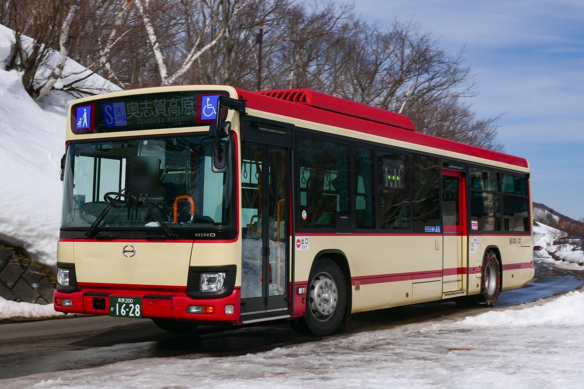 雪牆旁的山路上一輛紅色與米色的長電路線巴士，車頭顯示「S スキーシャトル 奥志賀高原」
