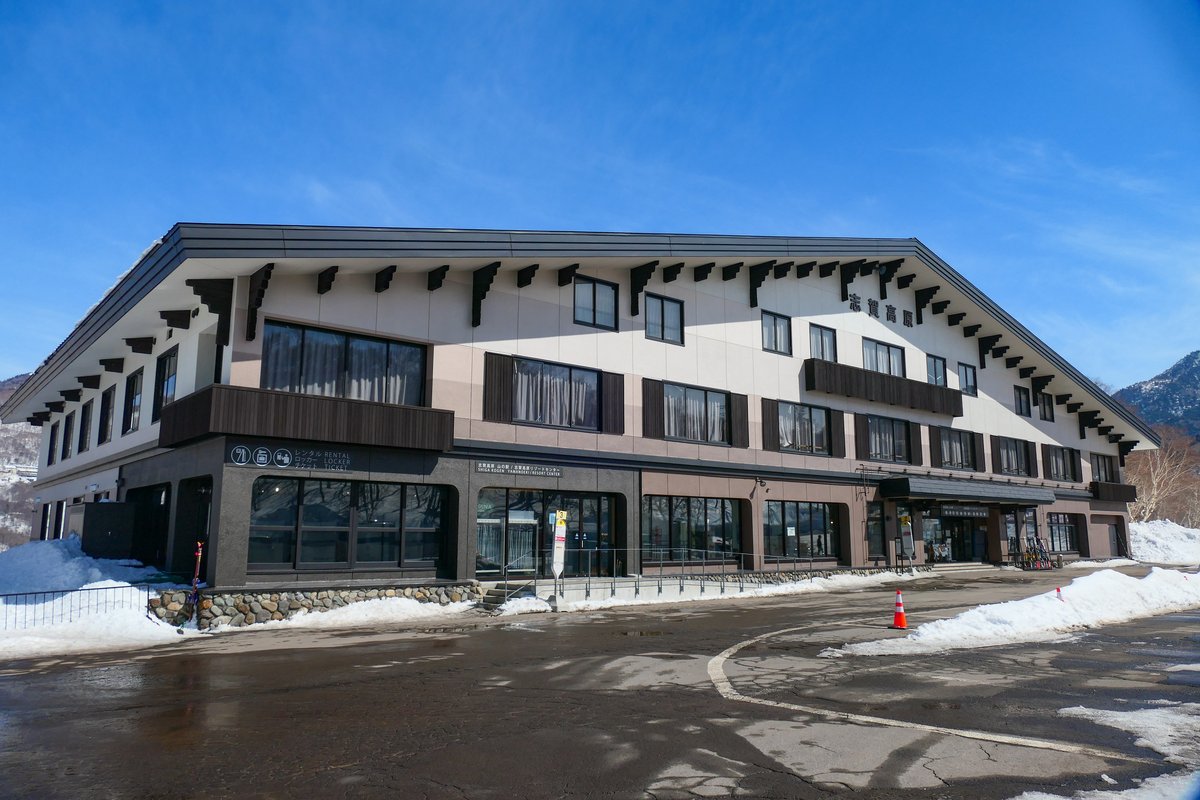 積雪路旁一棟三層的山屋風格建築，屋簷下寫「志賀高原」，一樓入口前立著一支巴士站牌