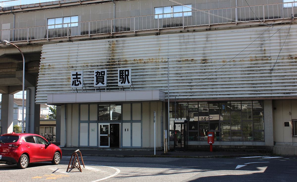 2020 年 10 月的 JR 志賀站站房：高架軌道下的米色建築，外牆掛著「志賀駅」三個大字，入口是玻璃門，門前有紅色郵筒，左側停著一輛紅色小客車