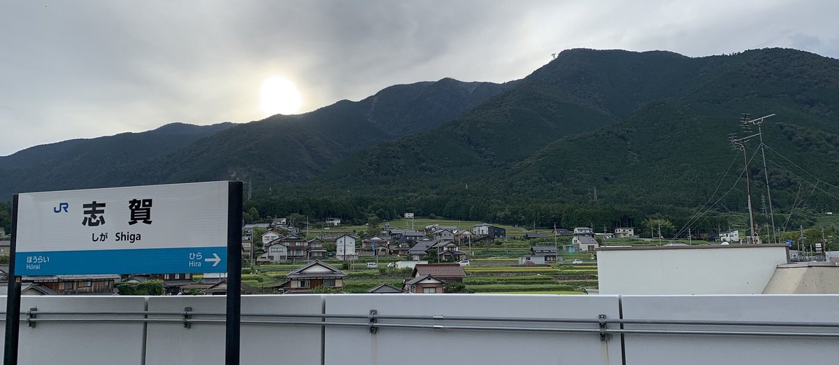 2023 年 9 月 JR 志賀站月台的站名牌，寫著志賀、しが、Shiga，前後站是蓬萊與比良；站名牌後方是聚落、梯田與比良山系的山