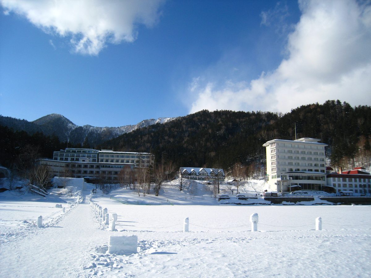 從結冰的然別湖湖面往岸上看：前景是冰上村的雪柱步道，岸邊右側是白色多層樓的ホテル風水，中間一棟半木造山形屋頂的小建築，左側是另一棟長形旅館，背後是覆雪的山