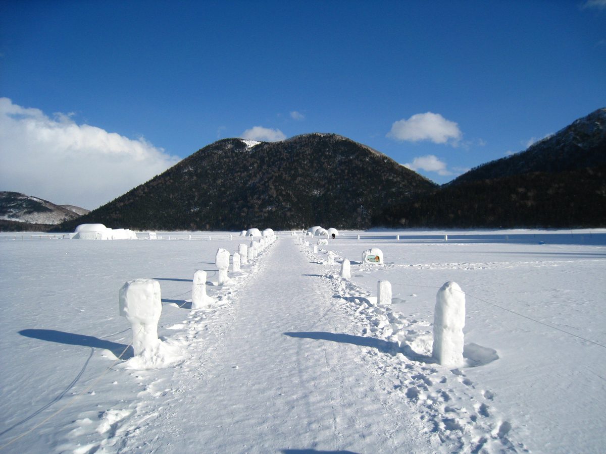 然別湖冰上村的湖面步道：兩側立著一排雪柱，步道一路通到遠處湖面上的幾棟冰屋，背後是整片覆著樹林的天望山與藍天