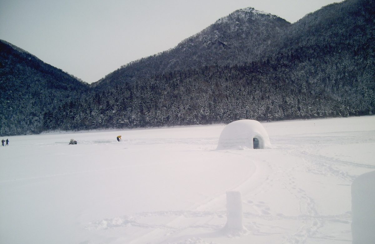 1 月下旬陰天的然別湖湖面，一大片雪地上只有一棟圓頂冰屋，遠處有幾個人和一台雪上車在湖上作業，背後是覆雪的針葉林山坡