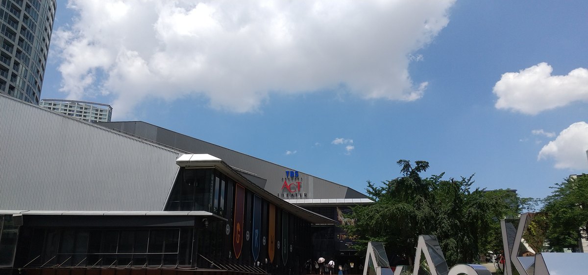 藍天白雲下的 TBS 赤坂ACT劇場:灰色金屬屋頂的低矮建築,正面上方有紅藍色的 TBS ACT THEATER 標誌,玻璃立面前垂掛一排四色長旗,右下角是銀色的 AKASAKA 立體字
