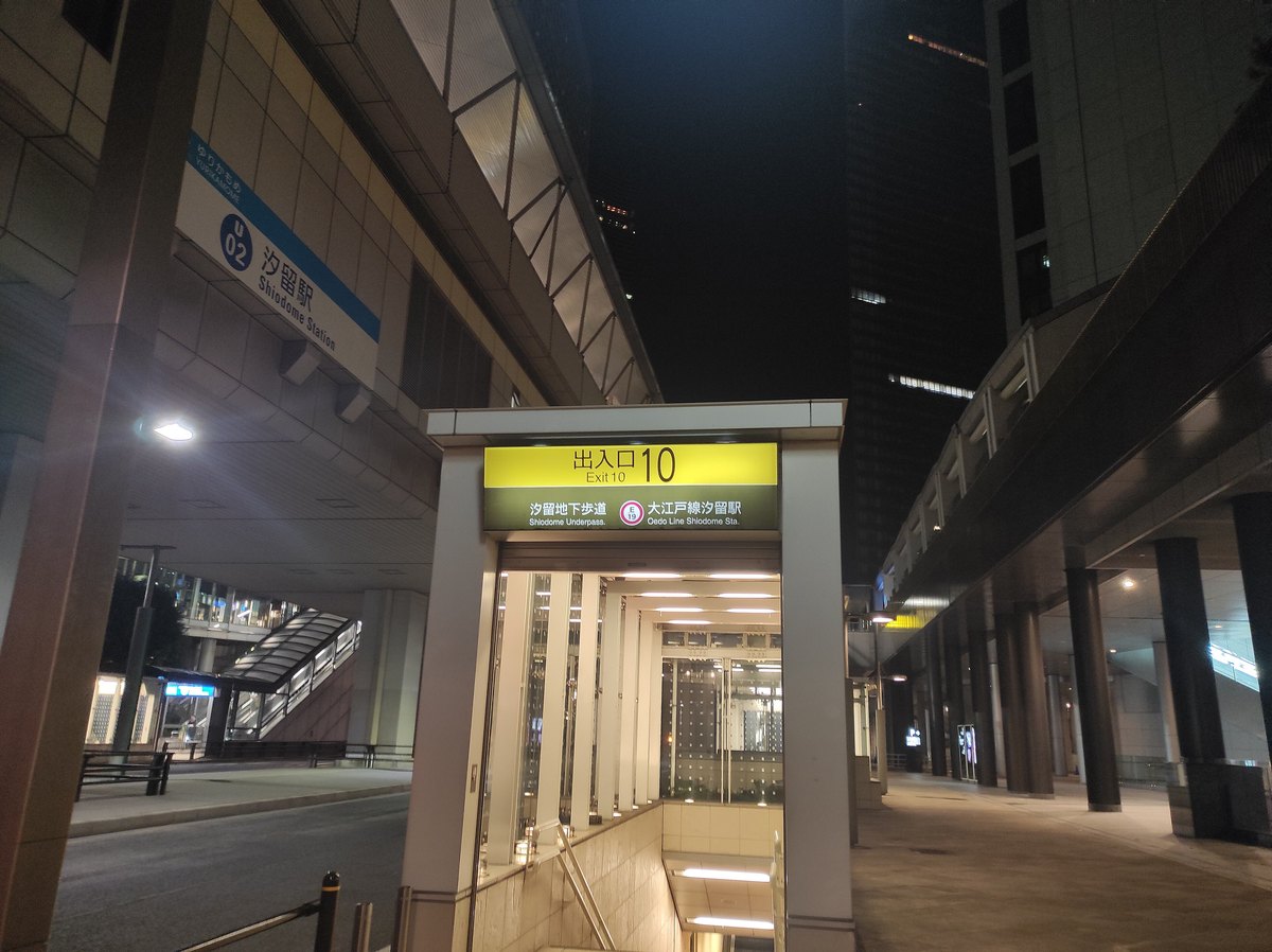 夜晚的汐留站 10 號出入口:白色方框的地下入口,上方黃色招牌寫著「出入口 10」與「大江戸線汐留駅」,左上方的高架下掛著百合海鷗號汐留站的藍色站名牌,右邊是高樓下的騎樓走廊