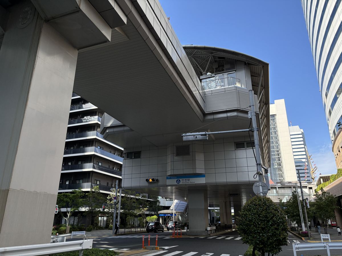百合海鷗號竹芝站從路口仰望:高架軌道從左上方延伸過來,車站本體是架在柱子上的灰白色方形量體,下方掛著「竹芝駅」藍色站名牌,馬路上有斑馬線與號誌,右邊一棟弧形玻璃高樓