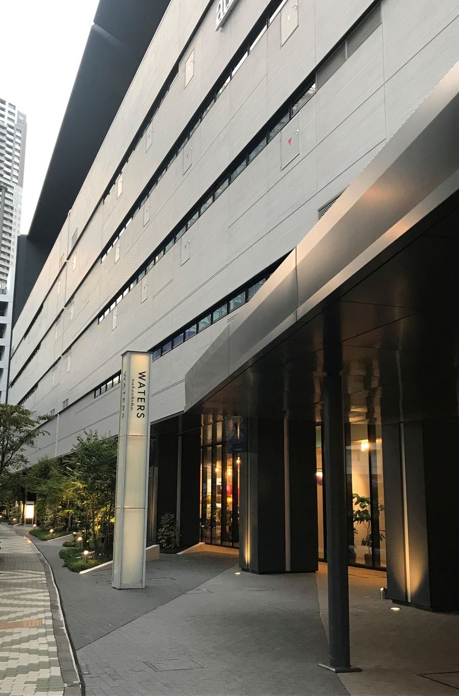 傍晚的竹芝劇場棟側面:一整面長條形的灰白色外牆、黑色屋簷伸出很遠,一樓是深色柱子撐起的騎樓,玻璃門內亮著暖色燈光,騎樓前立著寫有 WATERS takeshiba 的白色燈柱,左邊是植栽與石磚步道