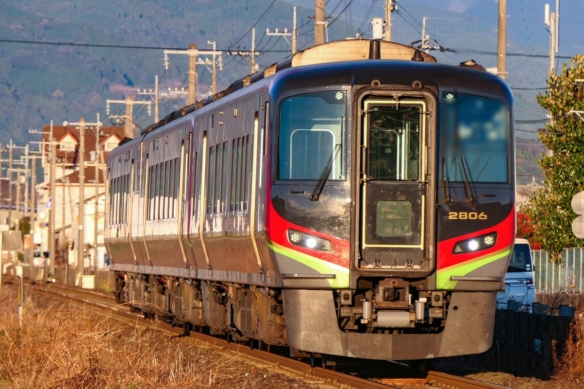 JR 四國 2700 系特急柴油車迎面駛來,深灰色車頭中央是貫通門,兩側有紅色與黃綠色飾帶,車號 2806,背景是電線桿、民宅與山坡,鐵道旁是枯黃的草