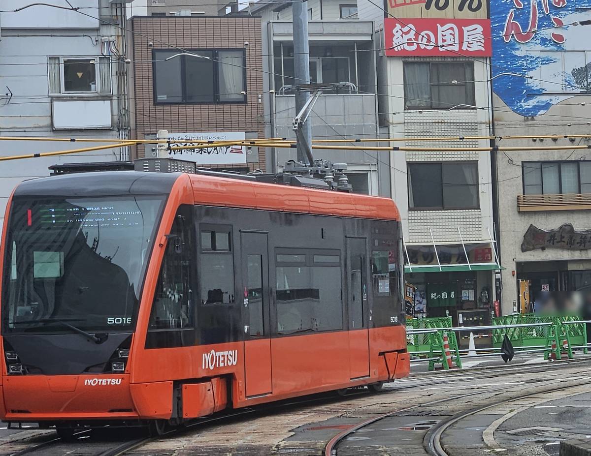 一輛橘色車身、黑色車窗帶的伊予鐵 5000 形低底盤路面電車在彎道上轉彎,車頭寫著「IYOTETSU」與車號 5016,後方是掛著「紀の国屋」紅色招牌的商店與路旁的綠色施工圍欄