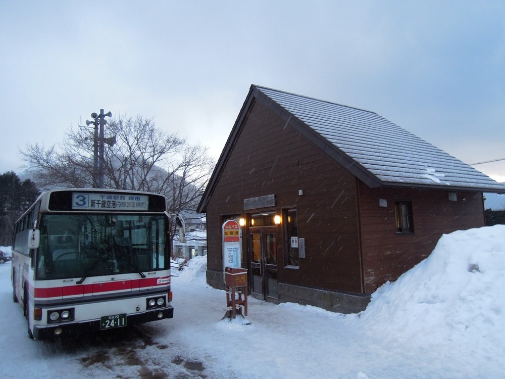 下雪的傍晚，北海道中央巴士「支笏湖」站：紅白塗裝的路線巴士停在積雪路邊，車頭顯示「3 千歳駅前経由 新千歳空港」，旁邊是咖啡色木造候車小屋和站牌，屋旁堆著雪