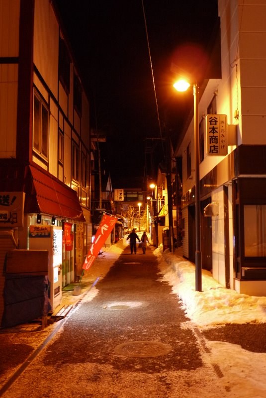 下過雪的夜晚，支笏湖溫泉街的一條窄街：路兩旁堆著雪，橘黃色的路燈照著路面，右邊建物掛著「谷本商店 酒・おみやげ」的招牌，左邊有紅色遮雨棚、自動販賣機和紅色旗幟，遠處兩個人並肩走著的背影