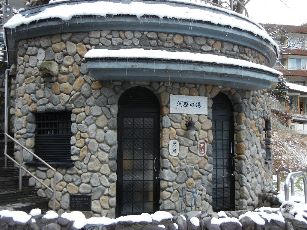 河原の湯：圓形的建築整面砌著河裡的卵石，中央掛著寫有「河原の湯」的石牌，左右兩扇拱形黑色鐵門分別標著男湯與女湯，屋簷上積著一層雪