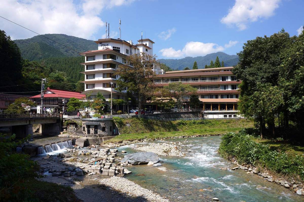 四万川的河床：白色卵石鋪滿溪底，水色偏淺綠，左邊是一座水泥道路橋，河對岸依序是紅屋頂的平房、一棟白色高樓旅館和一排長型旅館，後面整片是杉林山坡