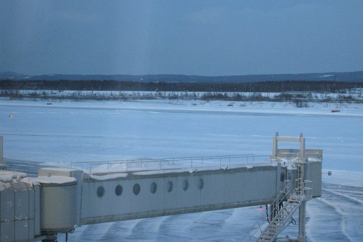 Snow-covered runway and apron at New Chitose Airport with a convoy of snow-clearing vehicles working the taxiway and an aircraft at the gate