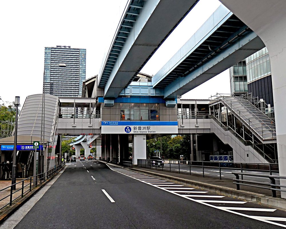 從馬路上看百合海鷗線新豐洲站:高架軌道橫過頭頂,軌道下方的站體掛著藍白色站名牌「U15 新豊洲駅 Shin-toyosu Station」,左右兩側各有一座通往地面的樓梯與電扶梯棟