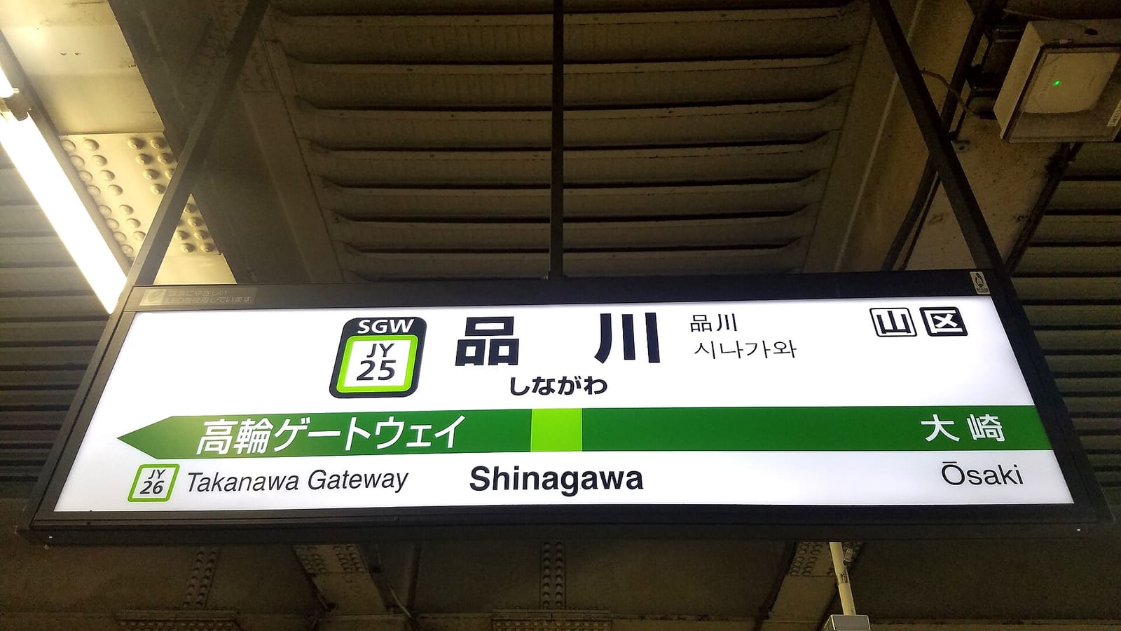 A JR platform nameboard at Shinagawa Station on the Yamanote Line, white with a black frame, reading Shinagawa in kanji, kana and roman letters, the green rounded-square station number JY 25 to its left, two small boxed characters in the top right corner, and a green band below marking the neighbouring stations Takanawa Gateway JY26 and Osaki