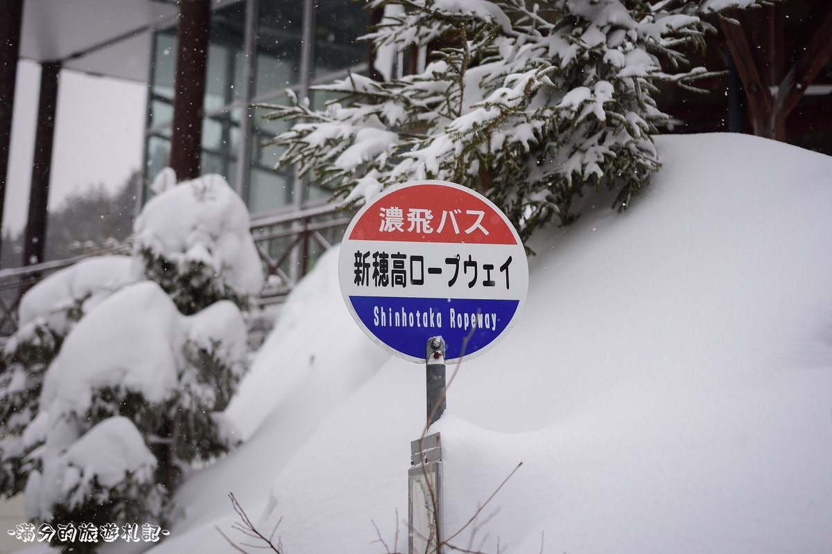 濃飛巴士「新穂高ロープウェイ」的圓形站牌立在及腰的積雪裡,背後是覆雪的樹叢,左側可以看到纜車站的玻璃帷幕與樓梯扶手